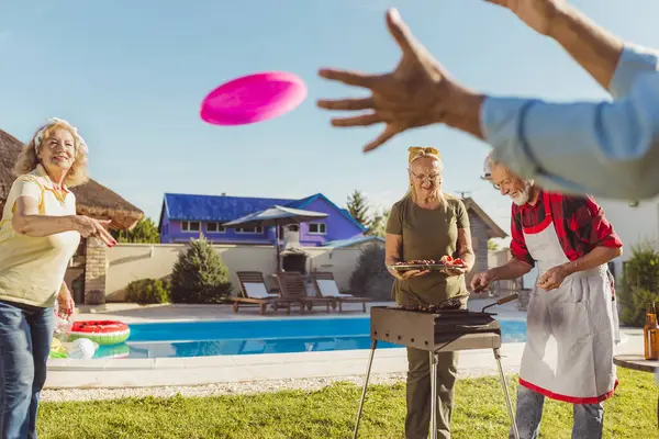 一群开朗活泼的老年人在后院举行烧烤会 烤肉和蔬菜 玩掷球接球游戏 — 图库照片