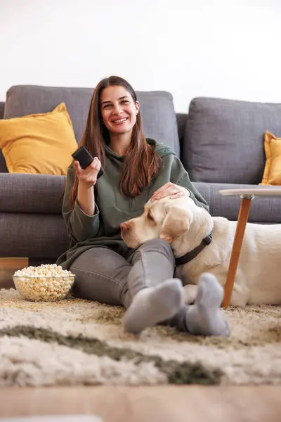 漂亮的年轻女人在家里休息 坐在客厅的地板上看电视 吃爆米花 而她的狗却躺在她身边 头枕在腿上 — 图库照片
