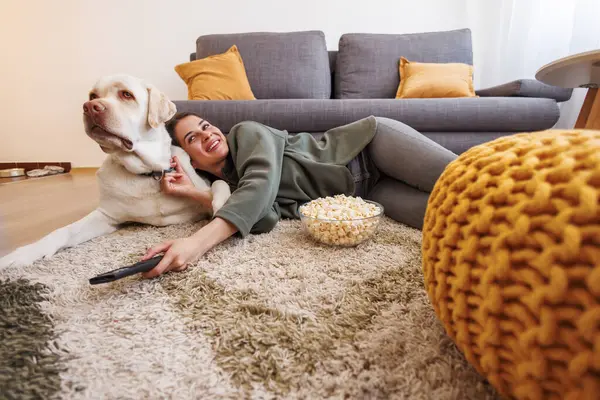 Beautiful young woman cuddling her pet dog while relaxing and enjoying leisure time at home, lying on living room floor eating popcorn and watching TV
