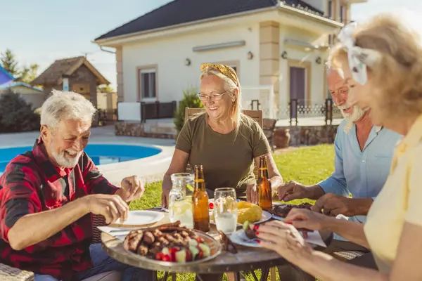 Group of senior people eating lunch in the backyard by the swimming pool, gathered around the table, eating, drinking and enjoying sunny summer day outdoors