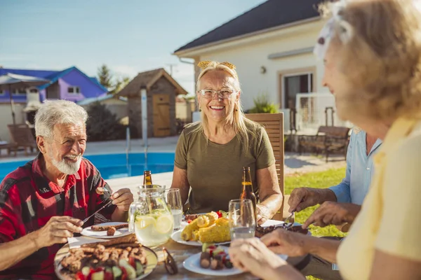 Group of senior friends eating lunch in the backyard by the swimming pool, gathered around the table, eating, drinking and relaxing on a sunny summer day outdoors
