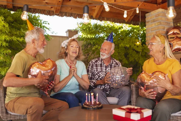 Group of cheerful senior friends celebrating friends birthday, wearing party hats and holding balloons while birthday girl is making a wish before blowing out candles on a cake