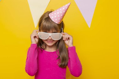 Küçük güzel bir kızın portresi parti gözlüğü ve doğum günü şapkası takıyor, doğum gününü kutluyor, sarı renkli arka planda izole edilmiş.