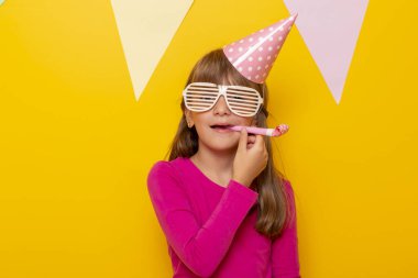 Güzel küçük kız doğum gününü kutluyor, parti gözlüğü takıyor ve parti düdüğü çalıyor, sarı renkli arka planda izole edilmiş.