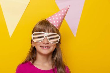 Küçük güzel bir kızın portresi parti gözlüğü ve doğum günü şapkası takıyor, doğum gününü kutluyor, sarı renkli arka planda izole edilmiş.