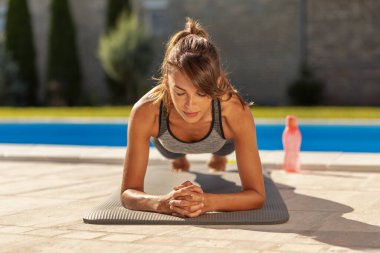 Genç bir kadın yüzme havuzunun yanında çalışırken ön kol egzersizi yapıyor.