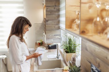 Güzel genç bir kadın mutfak lavabosunun yanında, mutfak beziyle bulaşıkları kurutuyor. 