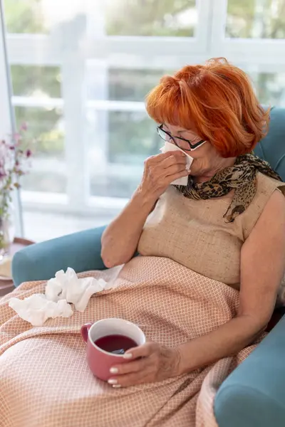 Sick elderly woman sitting in an armchair by the window covered with blanket, having flu and fever, blowing nose, sneezing and having a cup of hot tea