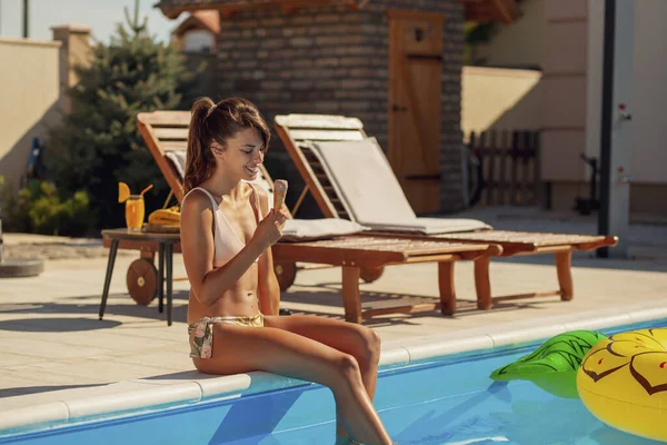 Beautiful young woman sitting at the edge of a swimming pool, relaxing on summer vacation, sunbathing and eating ice cream