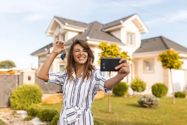 Beautiful young woman standing in front of her new house, taking a selfie using smart phone with the keys of her newly purchased property