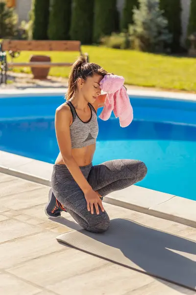Beautiful young woman wiping sweat using a towel after an outdoor workout in her backyard by the swimming pool