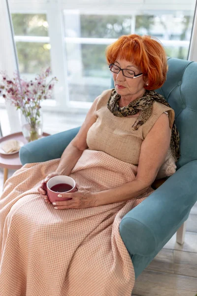 Elderly woman sitting in an armchair by the window covered with blanket, having fever and drinking cup of hot tea