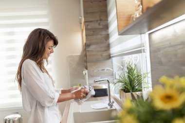 Güzel genç bir kadın mutfak lavabosunun yanında duruyor, mutfak bezini kullanarak bulaşıkları siliyor. 