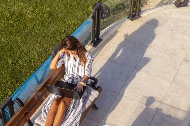 Evinin arka bahçesindeki terasta dizüstü bilgisayar kullanarak oturan serbest çalışan bir kadının yüksek açılı görüntüsü.