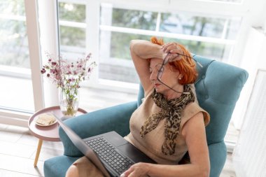 Kucağında dizüstü bilgisayarla koltukta oturan yorgun, yaşlı bir kadın. Alnında el ele tutuşuyor, sağlık sorunları yaşıyor.