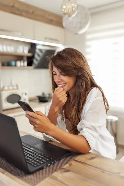 キッチンカウンターに座っている美しい若い女性 自宅から遠隔操作しながらスマートフォンとラップトップコンピュータを使用して — ストック写真