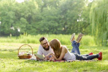Parkta piknik battaniyesinde yatan bir çift. Yanında meyve, yiyecek ve şarapla dolu bir piknik sepeti var. Tablet bilgisayarda internette sörf yapıyorlar.