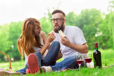 Parkta piknik battaniyesinde oturan bir çift. Adam nazikçe kızların saçlarına çiçek yerleştiriyor.