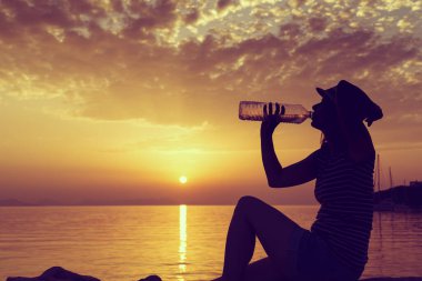 Kayalık sahilde oturan, su içen ve Yunanistan 'ın Syvota Körfezi' nde güzel bir günbatımının keyfini çıkaran genç bir kadın.