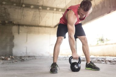 Genç, kaslı, atletik yapılı bir adam terk edilmiş bir binada ağırlık kaldırıyor.
