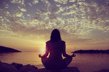 Yunanistan 'ın Syvota körfezindeki kayalık bir sahilde yoga yapan genç bir kadının silueti. Bulutlu gökyüzü, İyon denizi ve arka planda güzel bir günbatımı.