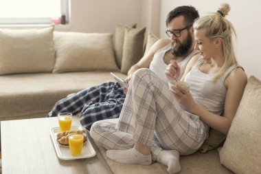 Aşık bir çift oturma odasındaki kanepede oturuyor, sabah kalktıktan sonra pijama giyiyor, tabet bilgisayarında haber okuyor ve kahvaltı yapıyor.