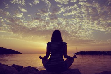 Yunanistan 'ın Syvota körfezindeki kayalık bir sahilde yoga yapan genç bir kadının silueti. Bulutlu gökyüzü, İyon denizi ve arka planda güzel bir günbatımı.