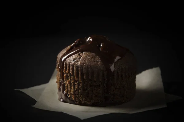 チョコレートマフィンは黒い背景にベーキングペーパーの上に置かれたチョコレートソースで覆われています — ストック写真
