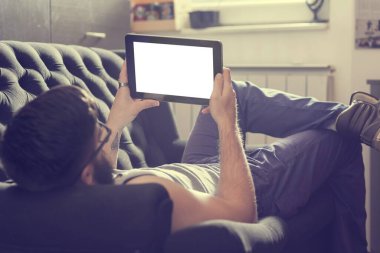 Oturma odasındaki kanepede uzanmış, tablet bilgisayarda internette sörf yapan bir erkek model.