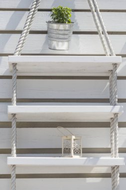 Dekoratif beyaz ahşap tahta bir duvarın kapağı halatlardan ve tahta tahtalardan yapılmış güzel raflar ve üzerine otlar konulmuş bir fener ve teneke kutu.