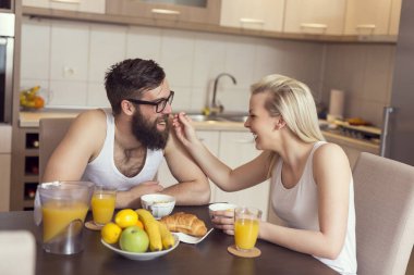 Aşık bir çift mutfak masasında oturuyor, birlikte kahvaltı yapıyor, kız adamı besliyor. Adama odaklan.
