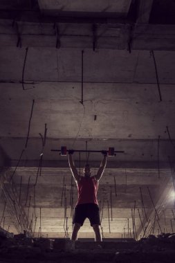 Genç kaslı adam terkedilmiş bir binada halter kaldırıyor.