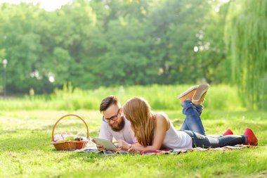 Birbirine aşık bir çift piknik sepetinin yanına yerleştirilmiş piknik sepetinde yatıp haberleri tablet bilgisayarda okuyor.