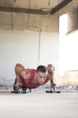 Kaslı, atletik, genç bir adam terkedilmiş bir harabe binada ek egzersiz malzemesi kullanarak şınav çekiyor.