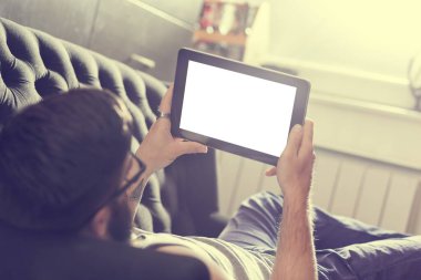 Oturma odasındaki kanepede uzanmış, tablet bilgisayarda internette sörf yapan bir erkek model.