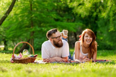 Parkta piknik battaniyesinde yatan bir çift. Yanında da meyve, yiyecek ve şarap dolu bir piknik sepeti var.