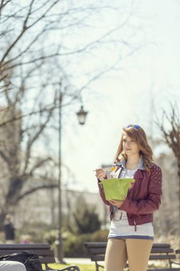 Genç, güzel esmer, güneşli bir günde parktaki ahşap bankta oturmuş Çin yemeği yiyor.