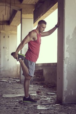 Kaslı, atletik yapılı, spor yapmadan önce harabe bir binada uzanan ve ayakta duran genç bir adam.