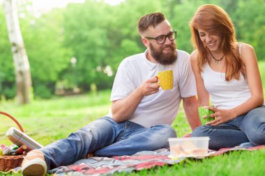 Genç aşık çift parkta ekose bir piknikte oturmuş kahve içiyor ve doğada günlerinin tadını çıkarıyorlar.