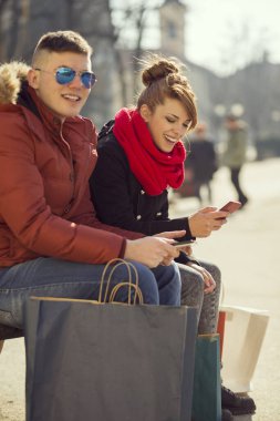 Parktaki ahşap bankta oturmuş internette sörf yapan genç bir çift.