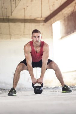 Genç, kaslı, atletik yapılı bir adam terk edilmiş bir binada ağırlık kaldırıyor.