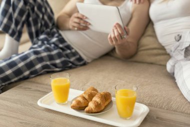 Aşık bir çift oturma odasındaki kanepede oturuyor, sabah kalktıktan sonra pijama giyiyor, tabet bilgisayarında haber okuyor ve kahvaltı yapıyor.