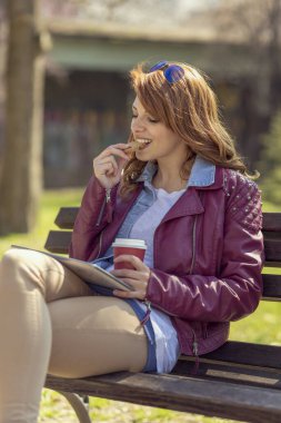 Genç, güzel esmer, parktaki ahşap bankta oturuyor, kahve içiyor ve güneşli bir günün tadını çıkarıyor.