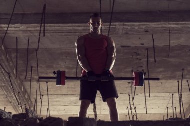 Genç kaslı adam terkedilmiş bir binada halter kaldırıyor.