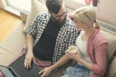 Birbirine aşık genç bir çift pencerenin yanındaki kanepede dinleniyor, internette dizüstü bilgisayarla geziniyor ve kahve içiyor. Pencere üzerinde parlaklık efekti