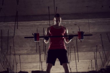 Genç kaslı sporcu halterle çalışıyor, terkedilmiş bir binada ağırlık kaldırıyor.