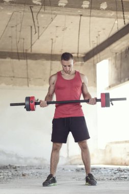 Terk edilmiş bir binada kaslı bir sporcu ağırlık kaldırıyor.