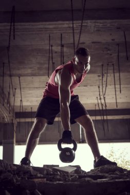 Genç, kaslı bir sporcunun, terkedilmiş bir binada ağırlık kaldırışının ön görüntüsü.