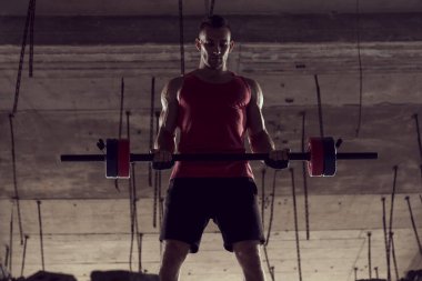 Genç kaslı sporcu halterle çalışıyor, terkedilmiş bir binada ağırlık kaldırıyor.