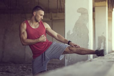 Kaslı, atletik yapılı, genç bir adam spor yapmadan önce uzanan harabe bir binanın duvarına bacağını kaldırıyor.